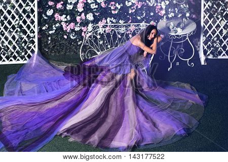 Beautiful brunette woman wearing long purple dress sitting on the bench in a flower garden