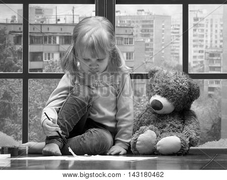 Little girl draws paints and brush. A huge window behind the glass city snow winter. toy teddy bear sitting next to. Black and white image