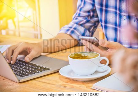 Close up Business man hand holding smart phone and use laptop in the coffee shop wood desk. vintage effect.