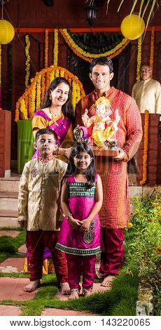 Smart Indian couple holding Ganesh idol or ganpati idol at entrance, welcoming lord ganesh on ganesh festival or chaturthi with kids and parents, happiness concept