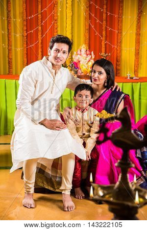 smart looking indian young couple in traditional wear sitting on sofa with boy child and ganesh idol or ganpati bappa in the background on ganesh festival or ganapati festival or utsav day