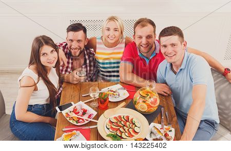 Group of happy young people posing at dinner table, party for friends indoors at cafe or home. Friendship, relax at holidays and week-end. Men and women sitting at table with various dishes.