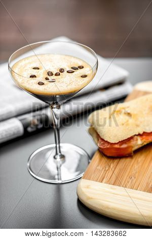 Shakerato drink in cocktail glass with panini and newspaper on the table. Italian lunch with traditional coffee and sandwich with prosciutto