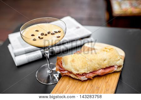 Shakerato drink in cocktail glass with panini and newspaper on the table. Italian lunch with traditional coffee and sandwich with prosciutto