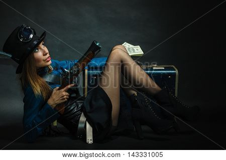 Fashionable subculture weapon concept. Steampunk girl with cash. Young gorgeous lady in victorian fashion lying on floor with banknotes briefcase and blunderbuss.