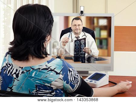 Experienced cardiologist sits face to face with online female patient which measures her blood pressure. Handsome virtual doctor attentively reviews brain x-ray results with her. Horizontal shot on blurry indoors background