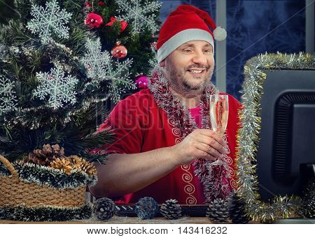 Cheerful middle-aged man in Santa Claus costume toasting glass of white sparkling wine in front of monitor. Bearded fat man sits at the desk surrounded tinsel garlands, balls, snowflakes, pine cones. He looks at the monitor his face illuminated by the scr