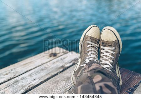 Man with crossed legs relaxing on riverbank pier casual young guy wearing sneakers sitting by the river in summer afternoon selective focus