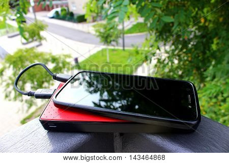 A black smartphone on a red powerbank lying on a fence in a residential district with a pond and lots of green vegetation