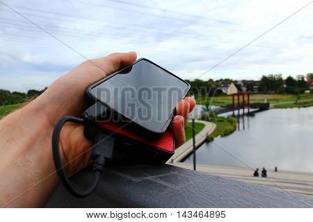 A black smartphone on a red powerbank lying on a fence in a residential district with a pond and lots of green vegetation