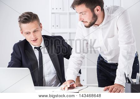 Businessmen at workplace. One is standing leaning to desk to write down information. Second is looking at computer screen and talking. Concept of cooperation.