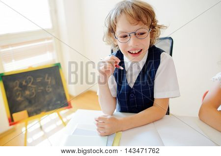 Cheerful little student bespectacled sits at school desk. It is a disciple of an elementary school. In a hand at boy ball pen. On a school desk copybooks lay. Near to a school desk a small blackboard.
