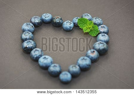 On a dark background blueberries ligned up in the shape of a heart and decorated with mint leaf.