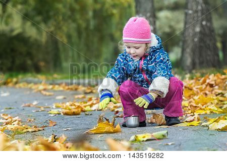 Two years old girl is stretching her hand to metal thermos flask cup hunkering at autumn foliage covering backdrop