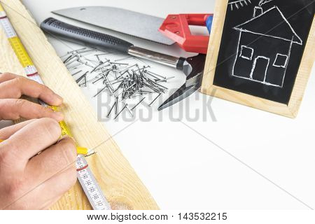 Builders hands measuring a wooden plank
Man's hands measuring a wooden board having his tools in the background and a funny sketch of a house on a blackboard. Concept about building your dream house.