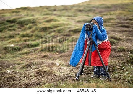 Little kid in a multi-colored big size windcheater is making a photo on the background of mountains. He is looking into the viewfinder of the camera which is on the tripod. Outdoors. Horizontal.
