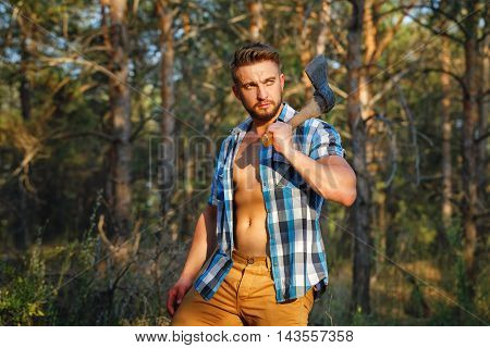 Lumberjack with an ax posing. Woodcutter in unbuttoned shirt in the coniferous forest. Felling trees. Logging. Manual labor. Brutal man.