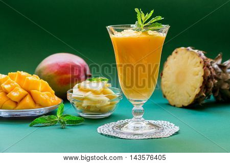 A glass of pineapple mango smoothie near ingredients on a green background. Pineapple mango smoothie. Horizontal. Daylight.
