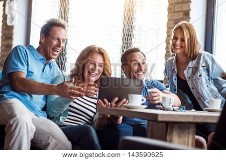 Laughable. Smiling and positive group of people using digital tablet together while drinking coffee in cafe