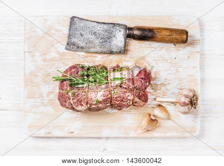 Raw uncooked roastbeef meat cut with rosemary, thyme and garlic and butcher knife on old white painted wooden background, top view, horizontal composition