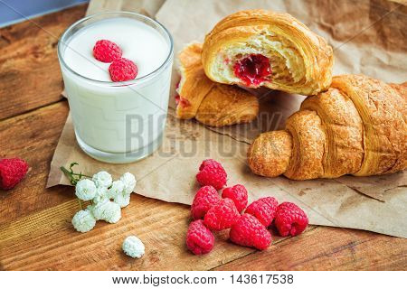 Cornetto with raspberry jam filling, glass of milk or yoghurt, fresh ripe berries and white flowers on the table, served for breakfast or snack