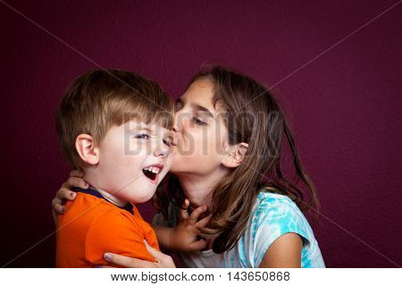 An older sister kisses her little brother on the cheek as he pushes her away with a disgusted look on his face.