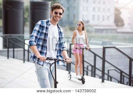 Rest with pleasure. Cheerful handsome smiling man riding a kick scooter and expressing joy with his friend
