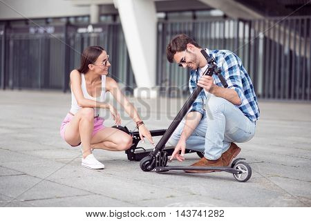 Share true emotions. Cheerful content friends sitting near kick scooter and smiling while resting