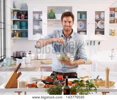 Handsome young man preparing salad at home in kitchen, smiling happy, looking at camera.