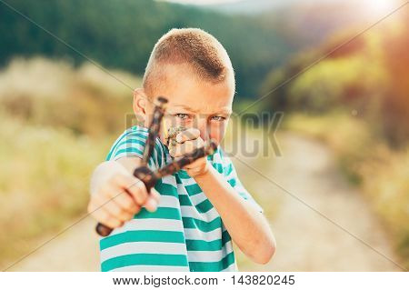 Naughty boy holding slingshot with stone. Little boy is playing in rural landscape.