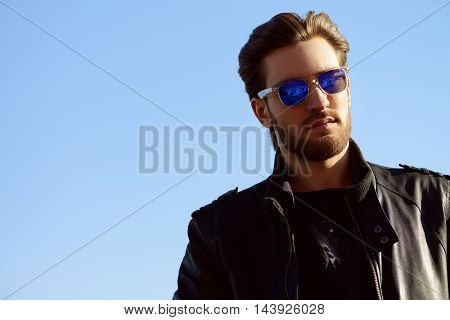 Confident handsome man in sunglasses and leather jacket over blue sky. Men's beauty, fashion. Outdoor portrait.