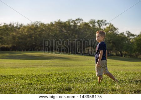 A cute kid boy standing alone in a large grassy meadow. Handsome child in the park looking into the distance. Boy playing on the grass. Weekend in the park. Leisure acitvities. Outdoor. Room for copy.