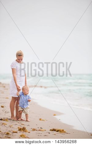 Adorable toddler boy standing by his young mother and looking at the ocean. Family by the sea. Summer vacations. First steps on the beach.Outdoor.