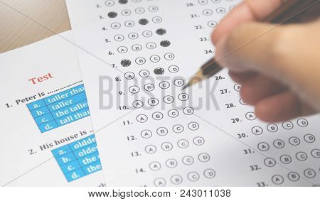 Multiple Choice Answer Sheet And English Test On Desk With Blur Hand Holding Pencil In Classroom