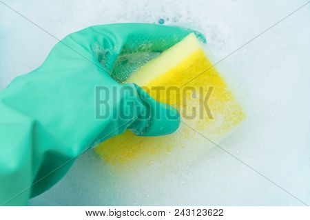 A Hand In A Turquoise Glove Holds A Sponge For Washing Dishes In A Copious Foam. Close-up