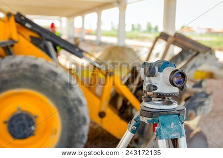 Surveyor Instrument For Measuring Level On Construction Site, Excavator Is In Background.