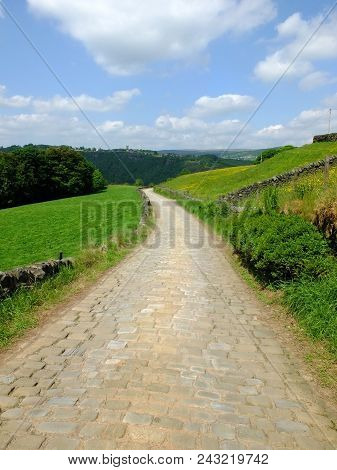 A Cobbled Country Lane Curving Downhill Into The Distant Wooded Valley Surrounded By Dry Stone Walls