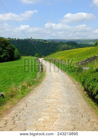A Cobbled Country Lane Curving Downhill Into The Distant Wooded Valley Surrounded By Dry Stone Walls