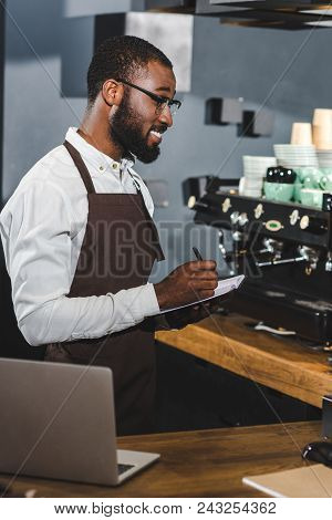 Smiling Young African American Barista In Eyeglasses Taking Notes While Woeking In Cafe