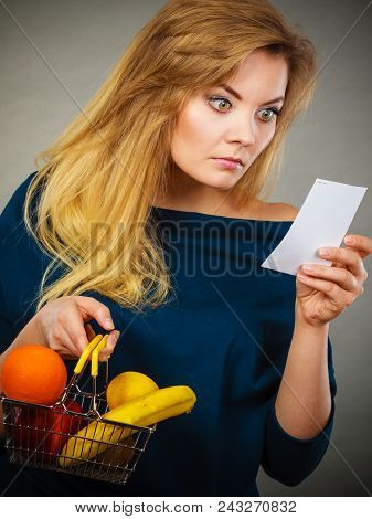 Worried Woman Holding Shopping Basket With Fruits Looking At Bill Receipt Being Scared Of Huge Price