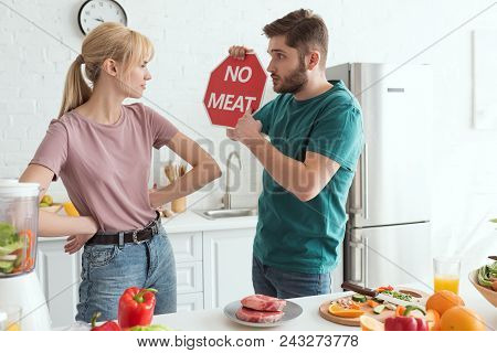 Vegan Couple With No Meat Sign At Table With Raw Meat And Vegetables In Kitchen At Home
