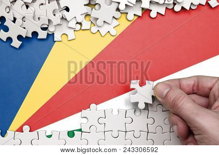 Seychelles Flag  Is Depicted On A Table On Which The Human Hand Folds A Puzzle Of White Color.
