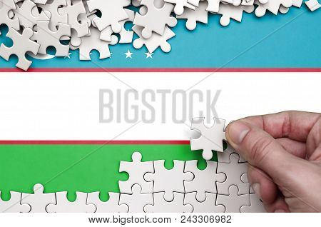 Uzbekistan Flag  Is Depicted On A Table On Which The Human Hand Folds A Puzzle Of White Color.