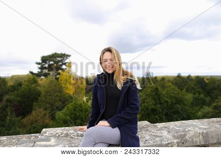 Young Female Person Sitting On Concrete Railing With Green Trees Background. Concept Of Architectura