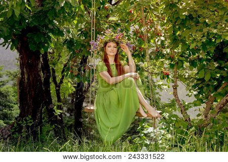 Young, Beautiful Woman In A Green Dress With A Wreath Of Wild Flowers Sits On A Homemade Swing In A 