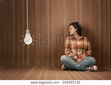 Young Woman Sits In  A Wooden Room And Looks At A Drawn Lightbulb.