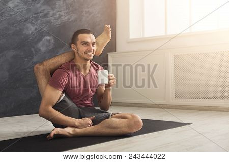 Young Sporty Man In Flexible Yoga Pose Drinking Tea, Sitting On Mat At Gym And Practicing Twisting E
