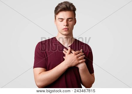 Shot Of Good Natured Young Hipster Male With Stylish Haircut, Keeps Hands On Heart, Demonstrates His