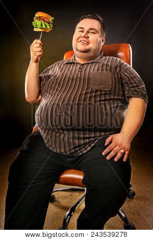 Diet failure of fat man eating fast food hamberger. Happy overweight person with wide-open mouth greedily eating huge hamburger. Health problems due to malnutrition. Business chairs concept.