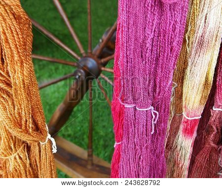 Colorful Hand-dyed Skeins Of Yarn Hang In Front Of A Wooden Spinning Wheel Against A Natural Backdro
